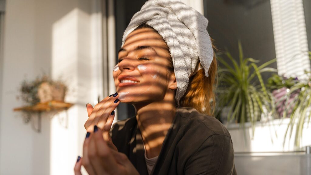 uma mulher com uma toalha na cabeça indicando que acabou de sair do banho, passando creme no rosto